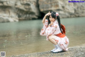 A woman in a pink kimono sitting on a rock by the water.